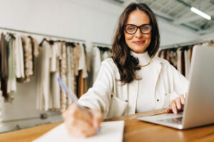 Portrait of a confident clothing store owner using a laptop as she writes down plans for her small business. Female entrepreneur using her creativity to manager and grow a successful fashion startup.